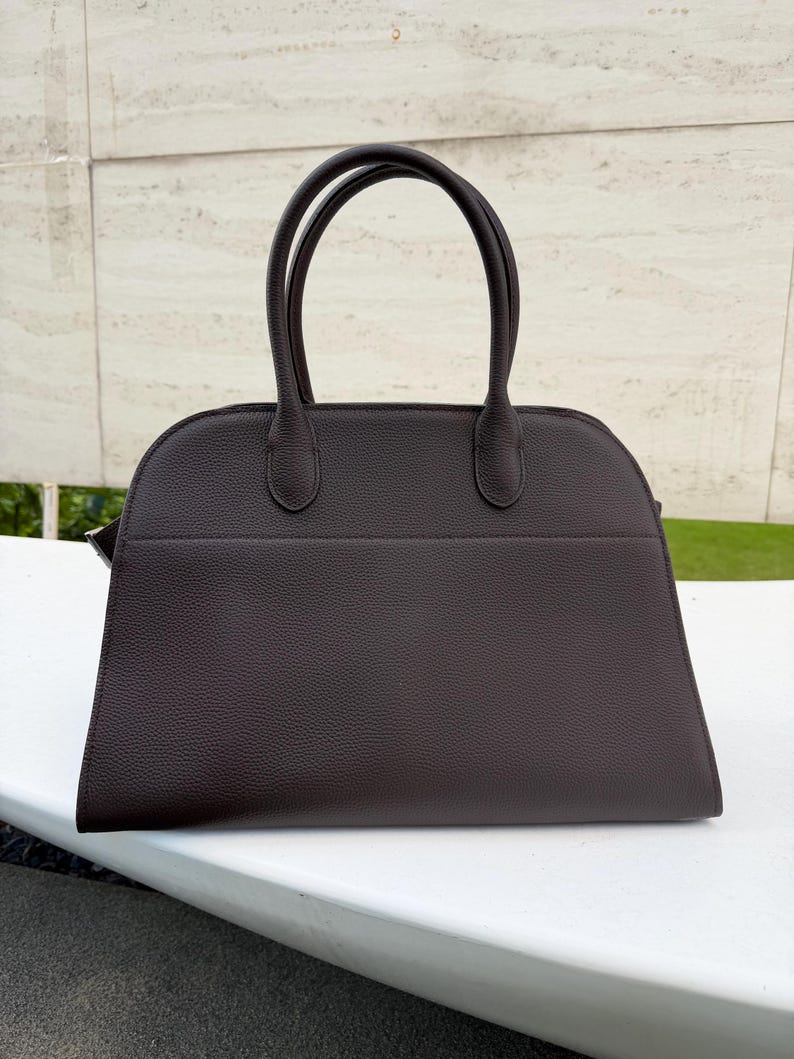 Puede incluir: Un bolso de mano marr&oacute;n oscuro con dos asas redondeadas. El bolso tiene una forma estructurada con una parte superior curva y una base rectangular. El material parece tener un acabado texturizado. El bolso est&aacute; sobre una superficie blanca.
