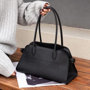 Puede incluir: Un bolso de mano negro con superficie texturizada, que cuenta con dos asas largas y delgadas y hebillas metálicas plateadas en las correas laterales. El bolso tiene un diseño estructurado con la parte superior curva y una costura horizontal.