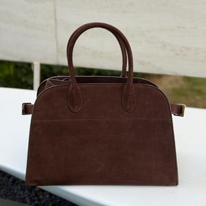Puede incluir: Un bolso de mano de ante marr&oacute;n con dos asas y forma rectangular. El bolso tiene una parte superior curvada y una base rectangular. El bolso tiene una hebilla dorada a cada lado. El bolso est&aacute; sobre una superficie blanca.