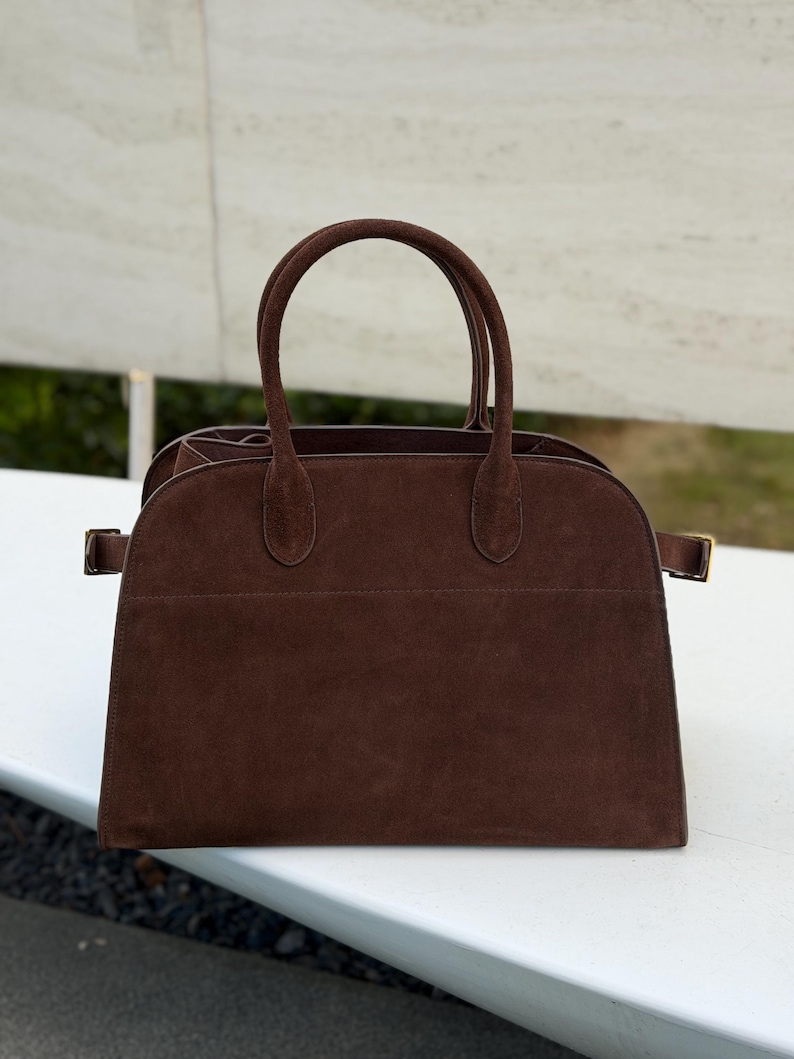 Puede incluir: Un bolso de mano de ante marr&oacute;n con dos asas y forma rectangular. El bolso tiene una parte superior curvada y una base rectangular. El bolso tiene una hebilla dorada a cada lado. El bolso est&aacute; sobre una superficie blanca.