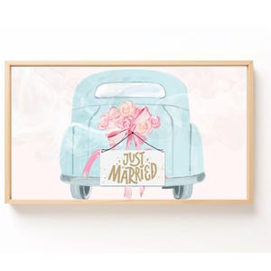 May include: Watercolor print of a light blue vintage truck with a pink bow and flowers in the back. A sign that reads "Just Married" hangs from the truck. The artwork is framed in a light wood frame.
