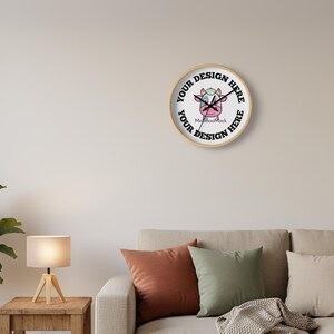 May include: A round wall clock with a light wood frame. The clock face features a cartoon cow design and the text "YOUR DESIGN HERE" in a circular pattern. The clock is hanging on a neutral-colored wall above a sofa.