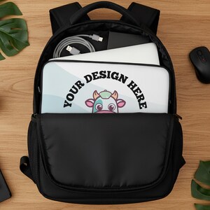 May include: Black backpack with a laptop sleeve featuring a cartoon cow design and the text "YOUR DESIGN HERE." The backpack is open, revealing a laptop, mouse, and charging cable. The background is a wooden surface.