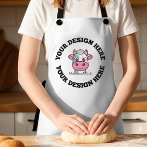 May include: White apron with black straps and a round graphic featuring a cartoon cow with the text "YOUR DESIGN HERE" encircling the cow. The apron is being worn while someone is kneading dough on a wooden surface.