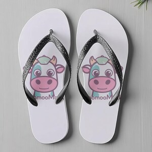 May include: A pair of white flip-flops with black straps. Each flip-flop has a cartoon cow face with the text "mooMoo". The cow has a pink nose, blue and green accents, and a smiling expression. Ideal for summer wear.