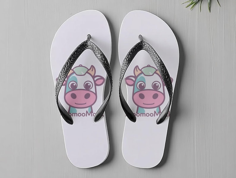 May include: A pair of white flip-flops with black straps. Each flip-flop has a cartoon cow face with the text "mooMoo". The cow has a pink nose, blue and green accents, and a smiling expression. Ideal for summer wear.