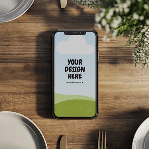 May include: A smartphone with the text "YOUR DESIGN HERE" on its screen, set against a sky and green hills design. The phone rests on a wooden table, with plates and cutlery arranged around it.