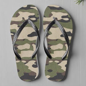 May include: A pair of camouflage-patterned flip-flops. The flip-flops have a green, brown, and beige camouflage design on the footbed. They feature black straps with a textured pattern. The background is a light grey surface.