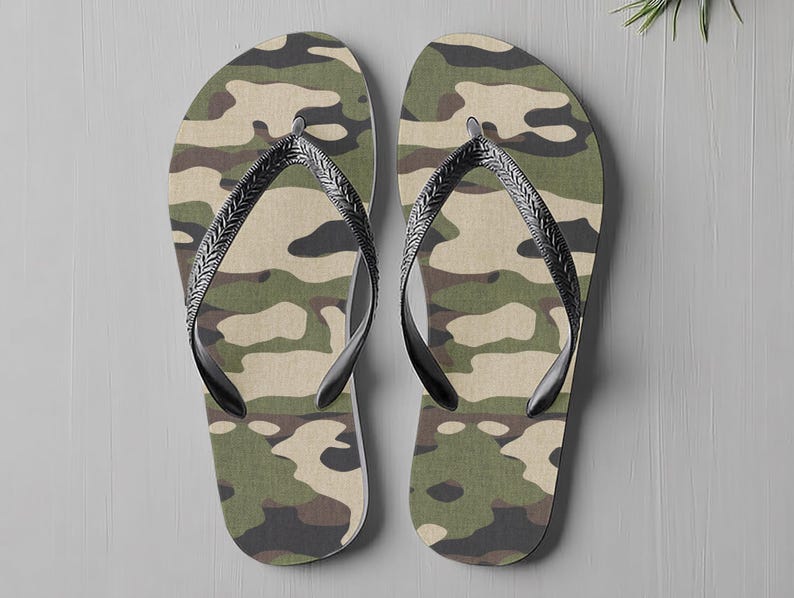 May include: A pair of camouflage-patterned flip-flops. The flip-flops have a green, brown, and beige camouflage design on the footbed. They feature black straps with a textured pattern. The background is a light grey surface.