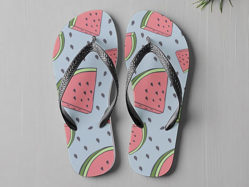 May include: A pair of flip-flops featuring a watermelon design on a light blue background. The watermelon slices are pink with green outlines and black seeds. The straps are black and silver.