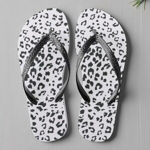 May include: A pair of white flip-flops featuring a black leopard print design. The straps are black and have a braided pattern. The flip-flops are shown on a grey surface.