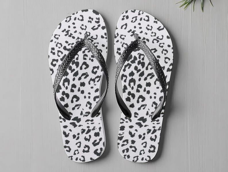 May include: A pair of white flip-flops featuring a black leopard print design. The straps are black and have a braided pattern. The flip-flops are shown on a grey surface.