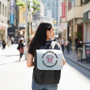 May include: Black backpack with a light blue flap featuring a cartoon cow design and the text "YOUR DESIGN HERE". The backpack has black straps and buckles. The person is walking down a city street.