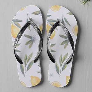 May include: Pair of white flip-flops featuring a lemon and leaf design. The sandals have black straps. The flip-flops are a summer accessory. The sandals are a summer accessory.