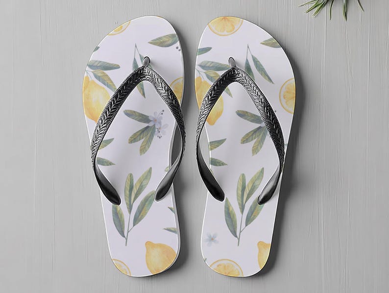 May include: Pair of white flip-flops featuring a lemon and leaf design. The sandals have black straps. The flip-flops are a summer accessory. The sandals are a summer accessory.