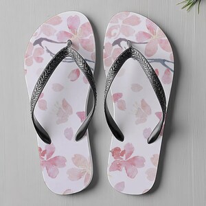 May include: A pair of white flip-flops with a pink floral pattern. The design features watercolour-style cherry blossoms and falling petals. The straps are black with a textured design. Ideal for a day at the beach or casual wear.