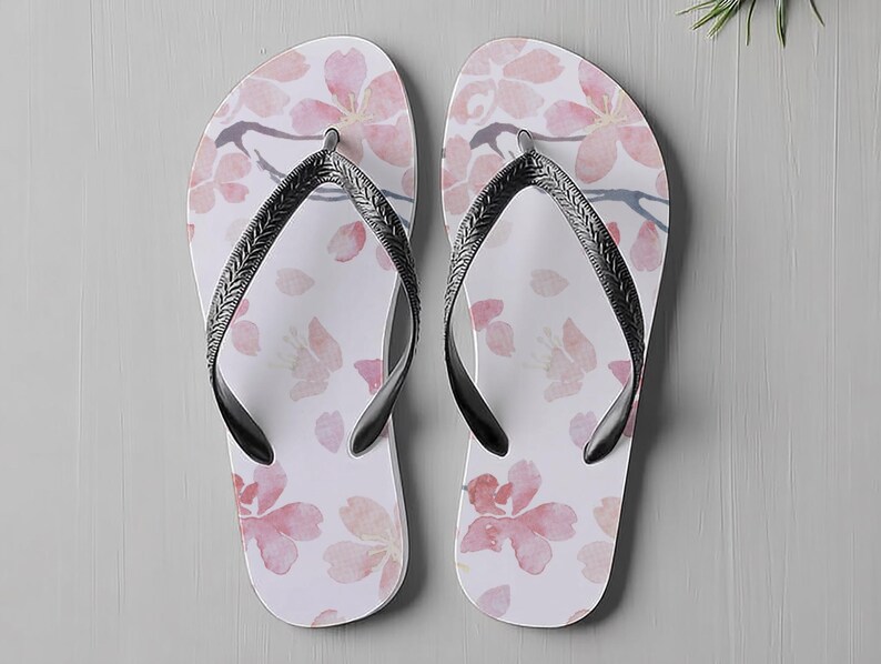 May include: A pair of white flip-flops with a pink floral pattern. The design features watercolour-style cherry blossoms and falling petals. The straps are black with a textured design. Ideal for a day at the beach or casual wear.