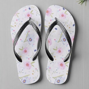 May include: A pair of white flip-flops with a floral pattern featuring pink and blue flowers. The flip-flops have black straps and are designed for casual wear. The sandals are a summer accessory.