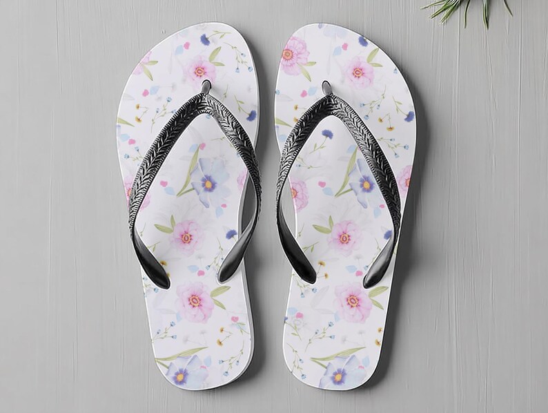 May include: A pair of white flip-flops with a floral pattern featuring pink and blue flowers. The flip-flops have black straps and are designed for casual wear. The sandals are a summer accessory.