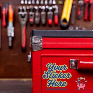 May include: A red toolbox with a black top and silver hardware. The toolbox has a handle and a sticker that says "Your Sticker Here" with a cartoon cow graphic. Tools hang in the background.