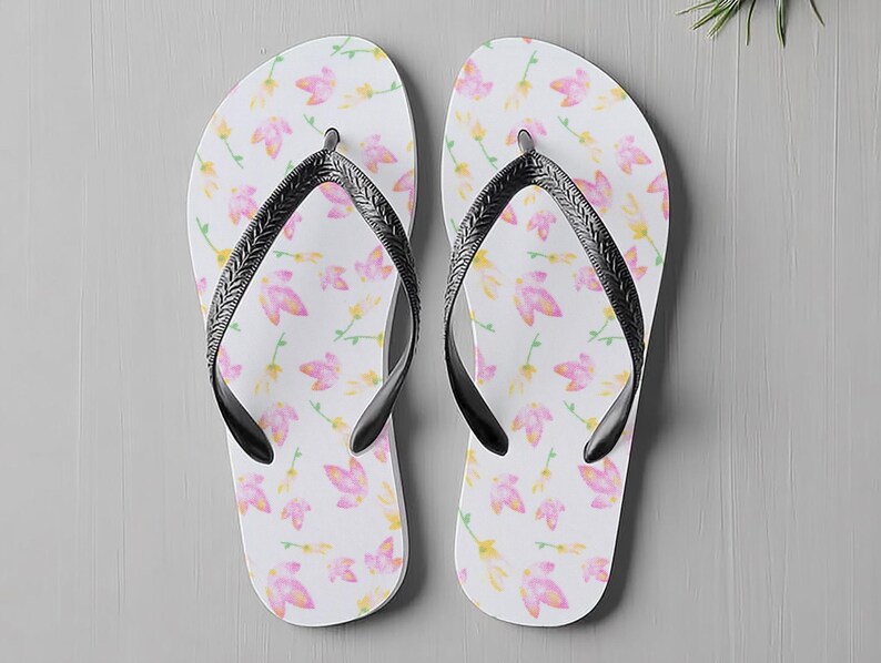 May include: White flip-flops featuring a pattern of pink and yellow flowers. The straps are black. These casual flip-flops are ideal for the beach or everyday wear, perfect for warm weather.