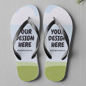 May include: A pair of white flip-flops with black straps. The flip-flops feature a design with a blue sky, clouds, and green grass. The text "OUR DESIGN HERE" and "MOOMOOMOCK" is printed on one flip-flop, and "YOU DESIGN HERE" and "MOOMOOMOCK" on the other.