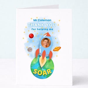May include: A white greeting card featuring a blue rocket ship with a circular photo of a child in the center. Below the rocket is a green and blue Earth graphic with the word SOAR in yellow. Text reads Mr Coleman THANK YOU for helping me.