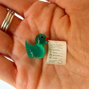 Könnte beinhalten: Eine kleine, durchscheinende grüne Entenfigur mit rotem Schnabel, die einen winzigen Papiernotiz hält. Die Notiz enthält Text und ein Datum. Die Ente liegt in der Handfläche, mit silbernen Ringen an den Fingern.