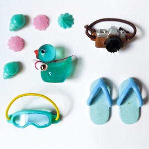 K&ouml;nnte beinhalten: Eine Sammlung von Miniatur-Accessoires zum Thema Strand, darunter eine Spielzeugkamera, Flip-Flops, eine Ente, eine Schwimmbrille sowie Muschel- und bl&uuml;tenf&ouml;rmige Dekorationen. Die Artikel sind in Blau-, Rosa- und Braunt&ouml;nen gehalten.