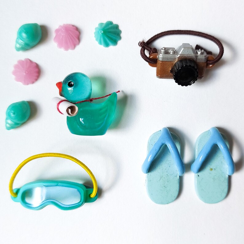 K&ouml;nnte beinhalten: Eine Sammlung von Miniatur-Accessoires zum Thema Strand, darunter eine Spielzeugkamera, Flip-Flops, eine Ente, eine Schwimmbrille sowie Muschel- und bl&uuml;tenf&ouml;rmige Dekorationen. Die Artikel sind in Blau-, Rosa- und Braunt&ouml;nen gehalten.