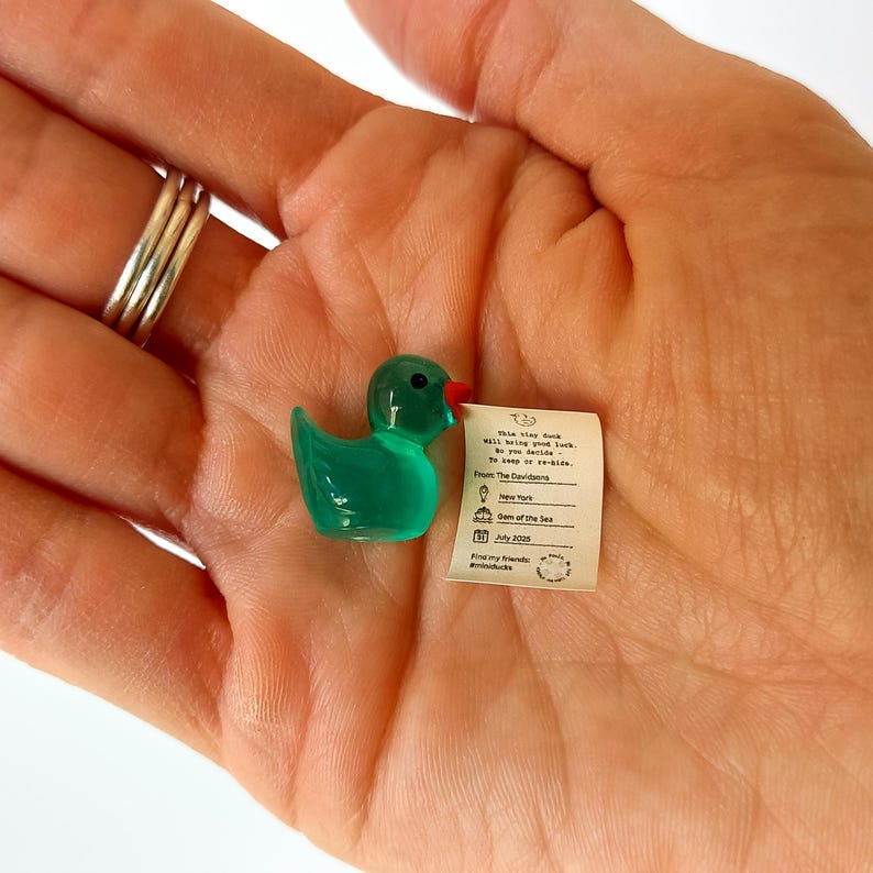 K&ouml;nnte beinhalten: Eine kleine, durchscheinende gr&uuml;ne Entenfigur mit rotem Schnabel, die einen winzigen Papiernotiz h&auml;lt. Die Notiz enth&auml;lt Text und ein Datum. Die Ente liegt in der Handfl&auml;che, mit silbernen Ringen an den Fingern.