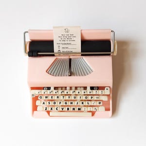 K&ouml;nnte beinhalten: Eine rosa Vintage-Schreibmaschine mit cremefarbenen Tasten und einer schwarzen Walze. Ein kleines Papieretikett ist angebracht, auf dem steht: "This tiny duck will bring good luck." Die Schreibmaschine hat einen Metallgriff und ein klassisches Design.