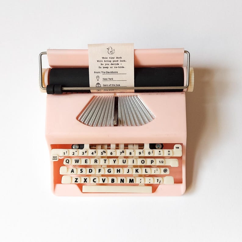 K&ouml;nnte beinhalten: Eine rosa Vintage-Schreibmaschine mit cremefarbenen Tasten und einer schwarzen Walze. Ein kleines Papieretikett ist angebracht, auf dem steht: "This tiny duck will bring good luck." Die Schreibmaschine hat einen Metallgriff und ein klassisches Design.