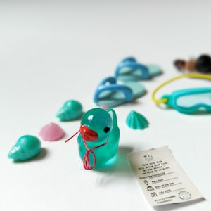 K&ouml;nnte beinhalten: Eine Sammlung von Miniatur-Spielzeugen zum Thema Strand, darunter eine t&uuml;rkisfarbene Ente mit rotem Schnabel, blaue Flip-Flops und t&uuml;rkisfarbene Muscheln. Eine kleine Karte mit Text ist ebenfalls vorhanden. Die Artikel sind auf einer wei&szlig;en Oberfl&auml;che angeordnet.