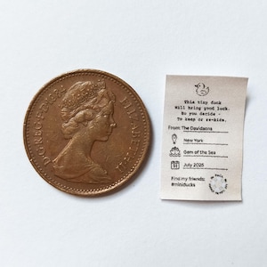 K&ouml;nnte beinhalten: Eine gro&szlig;e kupferfarbene M&uuml;nze neben einem kleinen, cremefarbenen Papier. Die M&uuml;nze zeigt das Profil einer Frau. Das Papier hat Text und eine Entenillustration, einschlie&szlig;lich der Worte "This tiny duck will bring good luck."