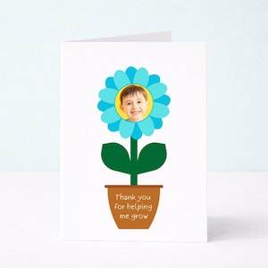 May include: A white greeting card featuring a graphic of a blue flower in a brown pot. The center of the flower contains a circular photograph of a smiling child. The text on the pot reads, Thank you for helping me grow, in white lettering.