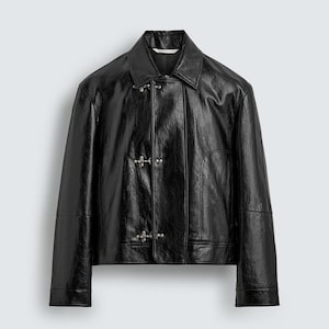May include: A black, glossy jacket with a collared neckline and three silver-toned clasps. The jacket has a slightly textured appearance and a boxy fit. The sleeves end at the wrist, and the jacket appears to be made of a leather-like material.