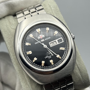 May include: A silver-toned Orient AAA Deluxe watch with a black face. The watch features a date display at the 3 o'clock position and luminous markers. The watch has a stainless steel band and is a vintage style.