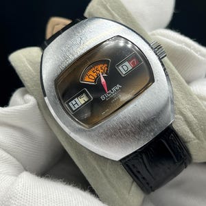 May include: Vintage Sicura automatic watch with a silver-tone case and a black leather band. The watch face features a brown dial with orange and white accents, including a date display and a unique time indicator. The watch is marked "Swiss Made".