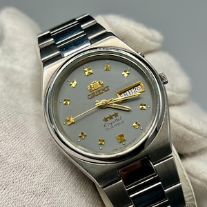 May include: A silver Orient Crystal 21 Jewels wristwatch with a grey face. The watch has gold hour markers and hands, and a day-date display. The watchband is made of silver metal.
