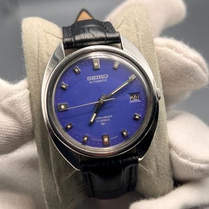 Puede incluir: Un reloj de pulsera automático Seiko vintage con una esfera azul vibrante y una pantalla de fecha. El reloj tiene una caja plateada, una correa de cuero negro y el texto "SEIKO AUTOMATIC WATER PROOF 17 JEWELS".