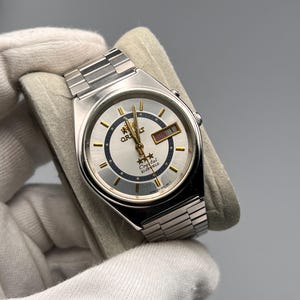 Op de afbeelding: Een zilverkleurig Orient Crystal horloge met een lichtblauwe wijzerplaat en een zilveren band. De wijzerplaat heeft goudkleurige wijzers en markeringen, met de dag en datum weergegeven. Het horloge wordt vastgehouden door een gehandschoende hand.