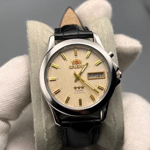 Peut inclure: Montre Orient argentée avec un cadran crème, des aiguilles et des index dorés, et un bracelet en cuir noir. Le cadran affiche la date et le logo Orient. La montre est présentée sur une surface grise.