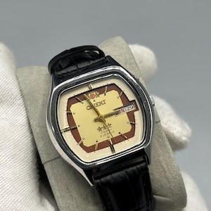 Könnte beinhalten: Eine Vintage-Orient-Uhr mit silbernem Gehäuse und schwarzem Lederarmband. Das Zifferblatt ist cremefarben mit braunen Akzenten, goldenen Zeigern und einer Tagesdatumsanzeige. Eine klassische Uhr.