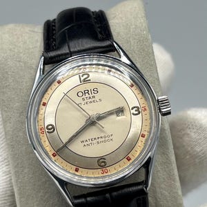 Może przedstawiać: Zegarek Oris Star w stylu vintage ze srebrną tarczą, czarnym skórzanym paskiem i kopertą ze stali nierdzewnej. Tarcza zegarka wyświetla godzinę za pomocą czerwonych i czarnych cyfr i wskazówek. Zegarek jest oznaczony "WATERPROOF ANTI-SHOCK".