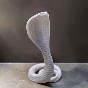 May include: A white cobra sculpture in a coiled position with its hood flared. The surface of the snake features detailed, textured scales across the entire body, including the underside of the hood and the coiled base, rendered in a matte finish.