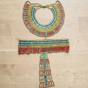May include: A beaded collar necklace and a matching wide belt with a dangling fringe panel. Both pieces feature horizontal rows of gold, turquoise, red, and black seed beads arranged in a geometric pattern with small turquoise-colored accents.