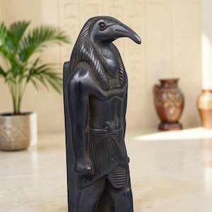 May include: A dark charcoal-colored statue of the Egyptian deity Thoth, depicted with the head of an ibis and a human body. The figure features detailed carvings of a traditional pleated kilt and a broad collar, standing in a rigid, upright pose.