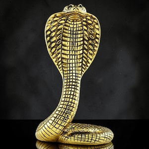 May include: A gold-coloured cobra figurine in a striking upright position with its hood flared. The sculpture features detailed, textured scales across its entire body, including the wide hood and the coiled base, with a metallic finish throughout.