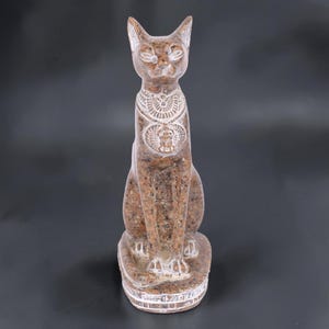 Può includere: Una statua in pietra maculata marrone di un gatto seduto con orecchie a punta. La figura presenta dettagli incisi in bianco attorno al collo, al petto e alla base, tra cui un motivo a scarabeo e fasce decorative che avvolgono il piedistallo.
